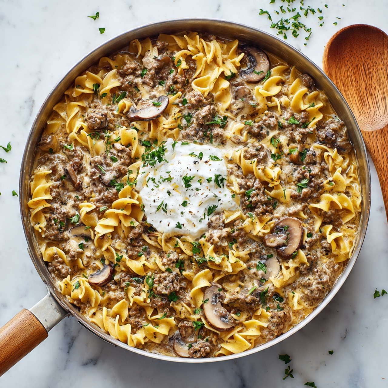 This image shows a round pan filled with a creamy noodle dish made of three layers: the first layer is twisted egg noodles in a light yellow color, spread evenly in the pan; the second layer consists of small crumbled cooked meat and sliced brown mushrooms scattered all around, blending with the noodles and covered with creamy sauce; the third layer is a dollop of white sour cream placed near the center, topped with a few small green parsley leaves and herbs sprinkled over the entire dish. The pan is on a white marbled surface with some green parsley around and a wooden spoon at the top right corner. photo taken with an iphone --ar 4:5 --v 7