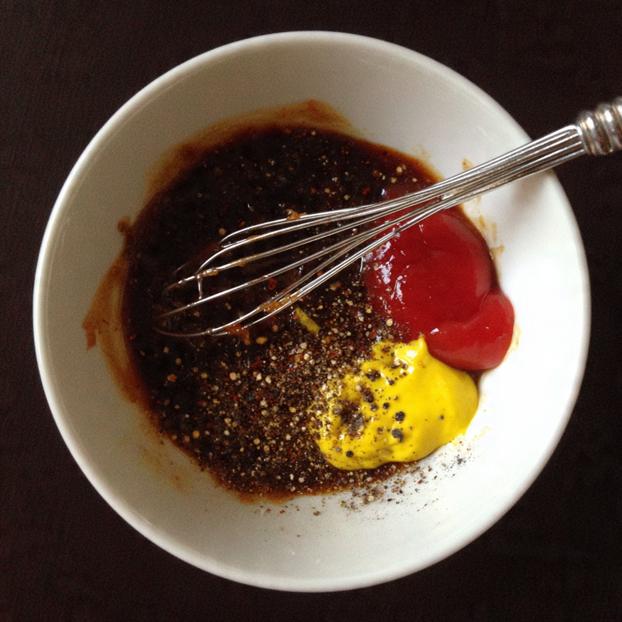A white bowl filled with a dark brown liquid sauce that has visible specks of black pepper and chili powder on top. There are dollops of red ketchup and bright yellow mustard on one side of the bowl. A silver metal whisk is inside the bowl, resting on the sauce and touching the sides. The bowl is placed on a white marbled surface. photo taken with an iphone --ar 4:5 --v 7