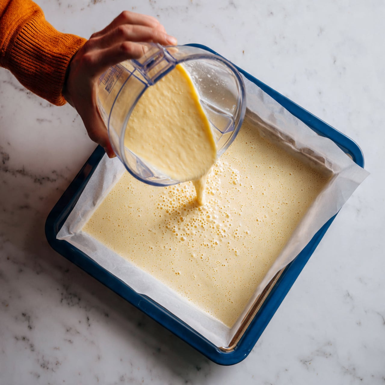 A smooth, pale yellow batter is being poured from a clear blender container onto a baking tray lined with white parchment paper. The batter spreads thin, with small bubbles forming on the surface, giving it a slightly frothy texture. The tray has blue edges and rests on a white marbled surface. A person wearing an orange sleeve is holding the blender container at the top right corner of the image. Photo taken with an iphone --ar 4:5 --v 7