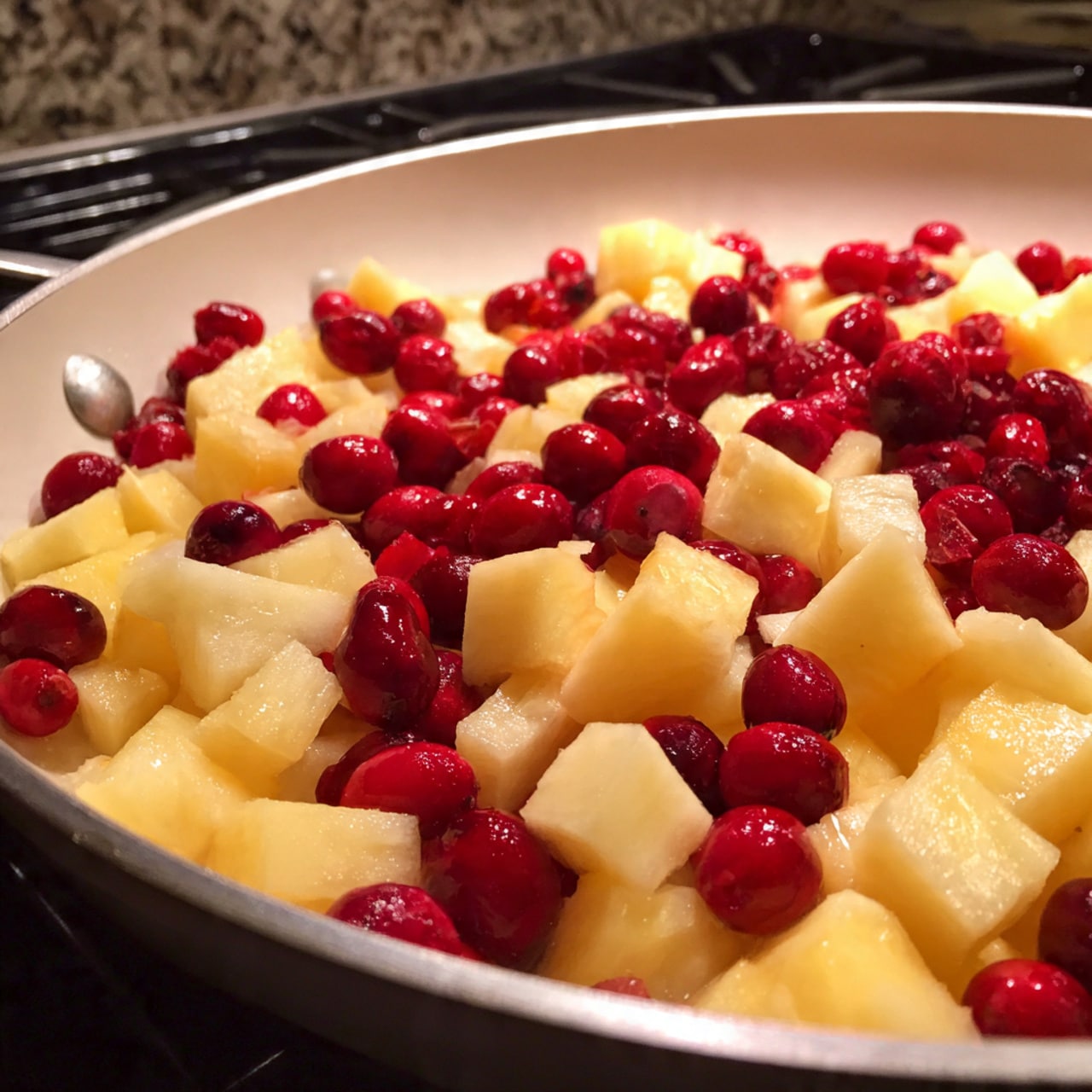 A white pan filled with a mixture of small, light yellow cubes of fruit and whole, vibrant red berries. The fruit cubes cover most of the pan's bottom, with the red berries scattered evenly among them. The pan is on a black electric stove, and the background has a white marbled texture. The colors are bright and fresh, showing a simple and natural fruit mixture photo taken with an iphone --ar 4:5 --v 7