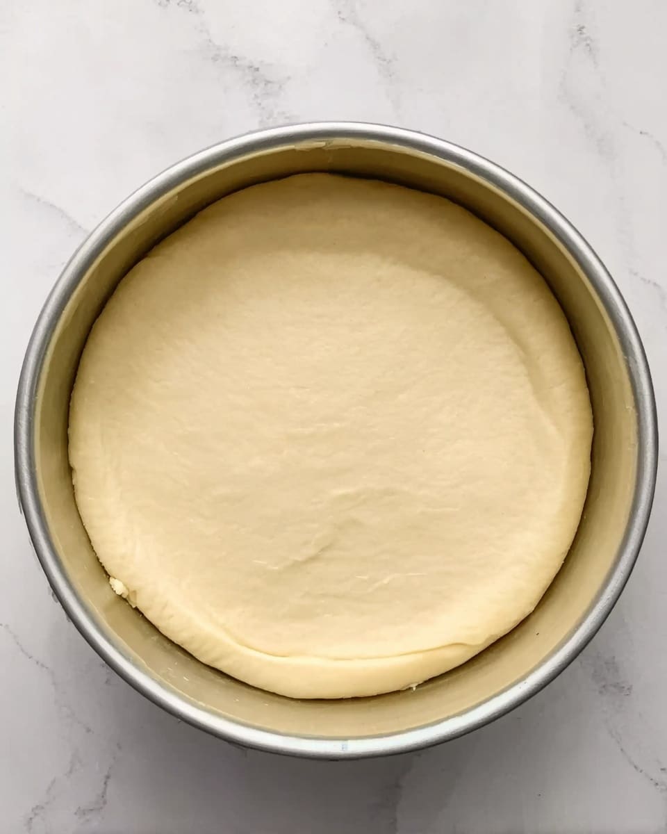 A round metal bowl with smooth, pale yellow dough evenly pressed flat inside it, filling the bottom and lightly rising along the sides. The dough looks soft and unbaked, with no cracks or uneven spots, resting on a white marbled surface. Photo taken with an iphone --ar 4:5 --v 7