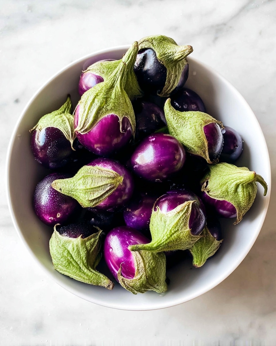 A white bowl filled with many small round eggplants with shiny deep purple skin and bright green caps. The eggplants are piled in layers, with the green caps pointing in different directions, showing their rough texture, while the smooth purple parts catch the light. The bowl sits on a white marbled surface, creating a clean and fresh look. photo taken with an iphone --ar 4:5 --v 7