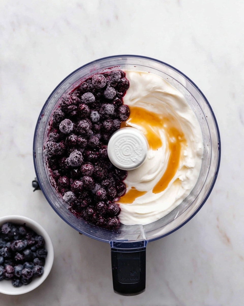 The image shows a clear food processor bowl with three layers inside. The left half is filled with dark purple frozen berries with a slightly frosty texture. The top right part has a smooth white layer of thick yogurt. Mixed in the yogurt on the right edge, there is a light amber drizzle of honey that blends gently into the white layer. The food processor bowl sits on a white marbled surface with a small white bowl of extra frozen berries placed beside it. Photo taken with an iphone --ar 4:5 --v 7