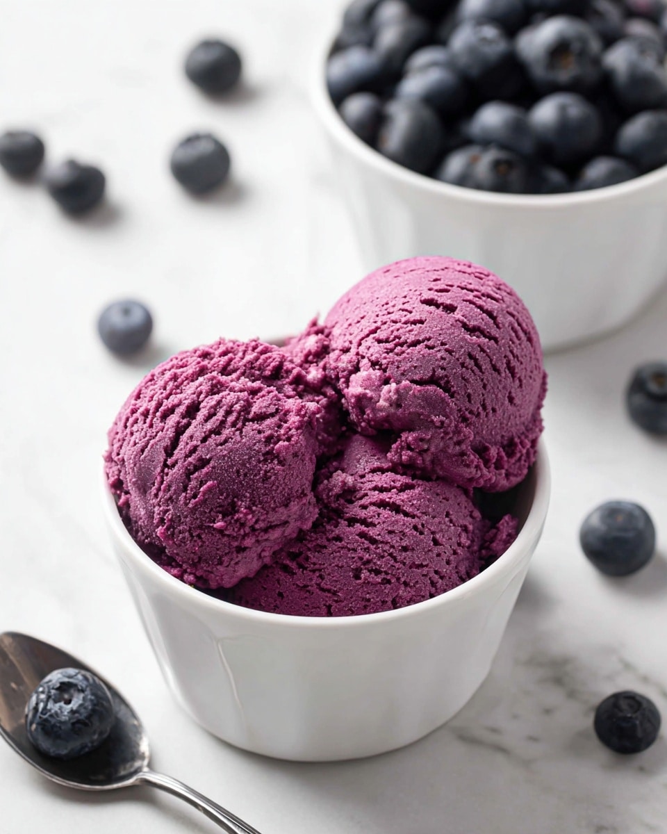 A white bowl filled with two scoops of textured, bright purple ice cream sits on a white marbled surface. The ice cream looks thick and slightly rough. In the background, another white bowl is filled with fresh, dark blue blueberries, with some blueberries scattered on the marble around the bowls. A silver spoon holds one large blueberry in the lower left corner of the image. photo taken with an iphone --ar 4:5 --v 7