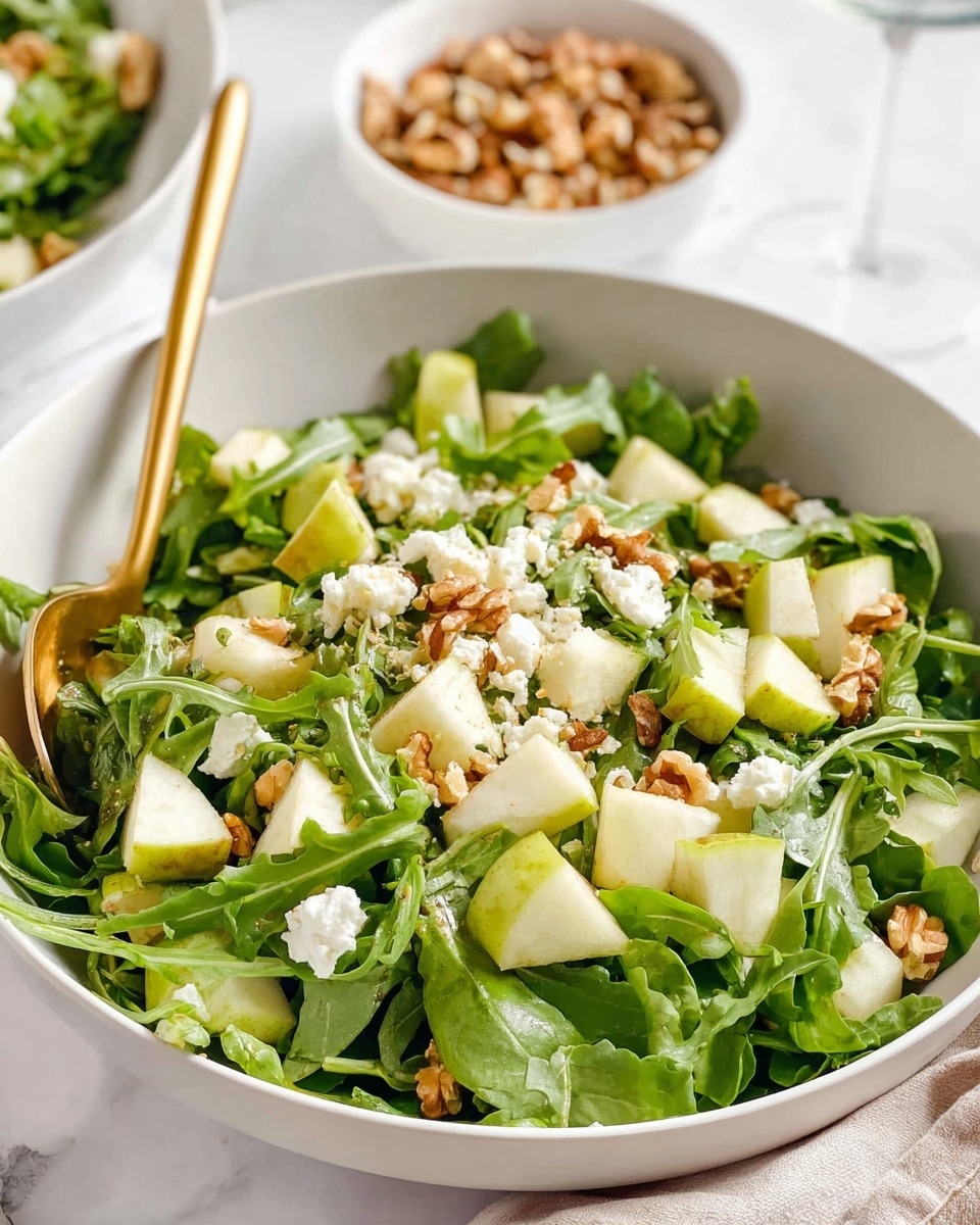 A white bowl filled with a fresh salad showing three main layers; the bottom layer is bright green arugula leaves, the middle layer has pale green and white cubes of pear, and the top layer is scattered with small white crumbles of cheese and light brown walnut pieces. A gold spoon rests inside the bowl on the left side, and in the background, there is a small white bowl filled with walnut halves. The whole scene is set on a white marbled surface, with soft natural light highlighting the fresh textures. photo taken with an iphone --ar 4:5 --v 7