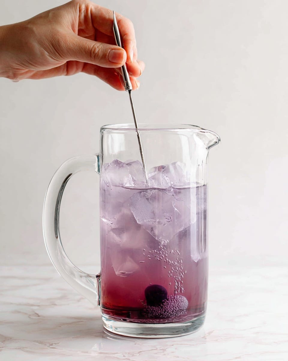 A clear glass pitcher filled halfway with light purple liquid and ice cubes inside is placed on a white marbled surface. A person's hand holds a long, thin metal stirrer dipping it into the liquid, causing small ripples and bubbles around a darker purple ingredient sinking at the bottom. The background is plain white, making the pitcher and the subtle color gradient of the drink the main focus. Photo taken with an iphone --ar 4:5 --v 7
