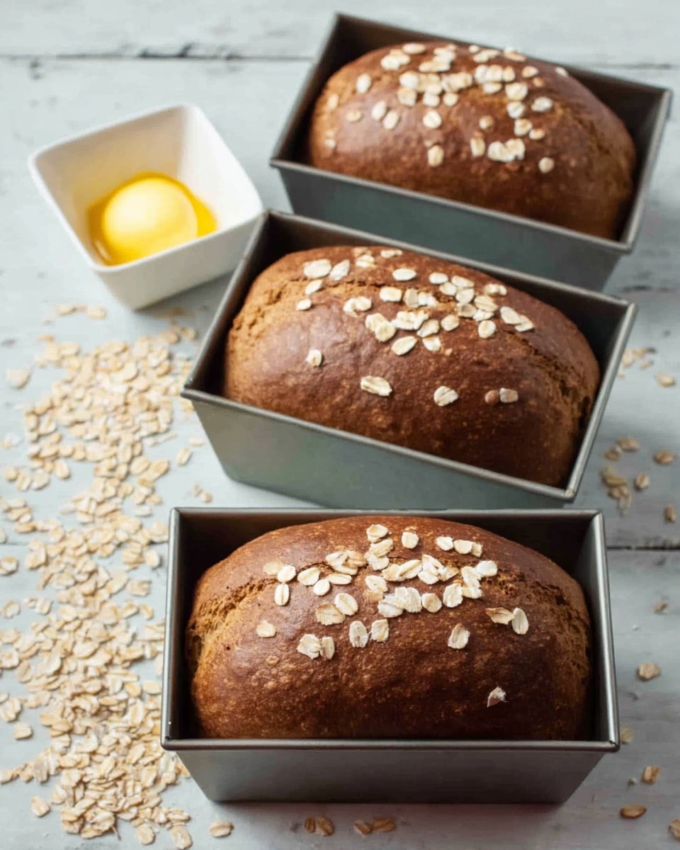 The image shows three freshly baked brown loaves of bread in small metal loaf pans, arranged in a triangular pattern on a white marbled surface. Each loaf is topped with scattered pale oat flakes. In front of the loaves, to the left, there is a small white square dish containing a dollop of soft yellow butter. Oat flakes are also scattered loosely around the loaves and butter dish. The photo is taken with an iphone --ar 4:5 --v 7