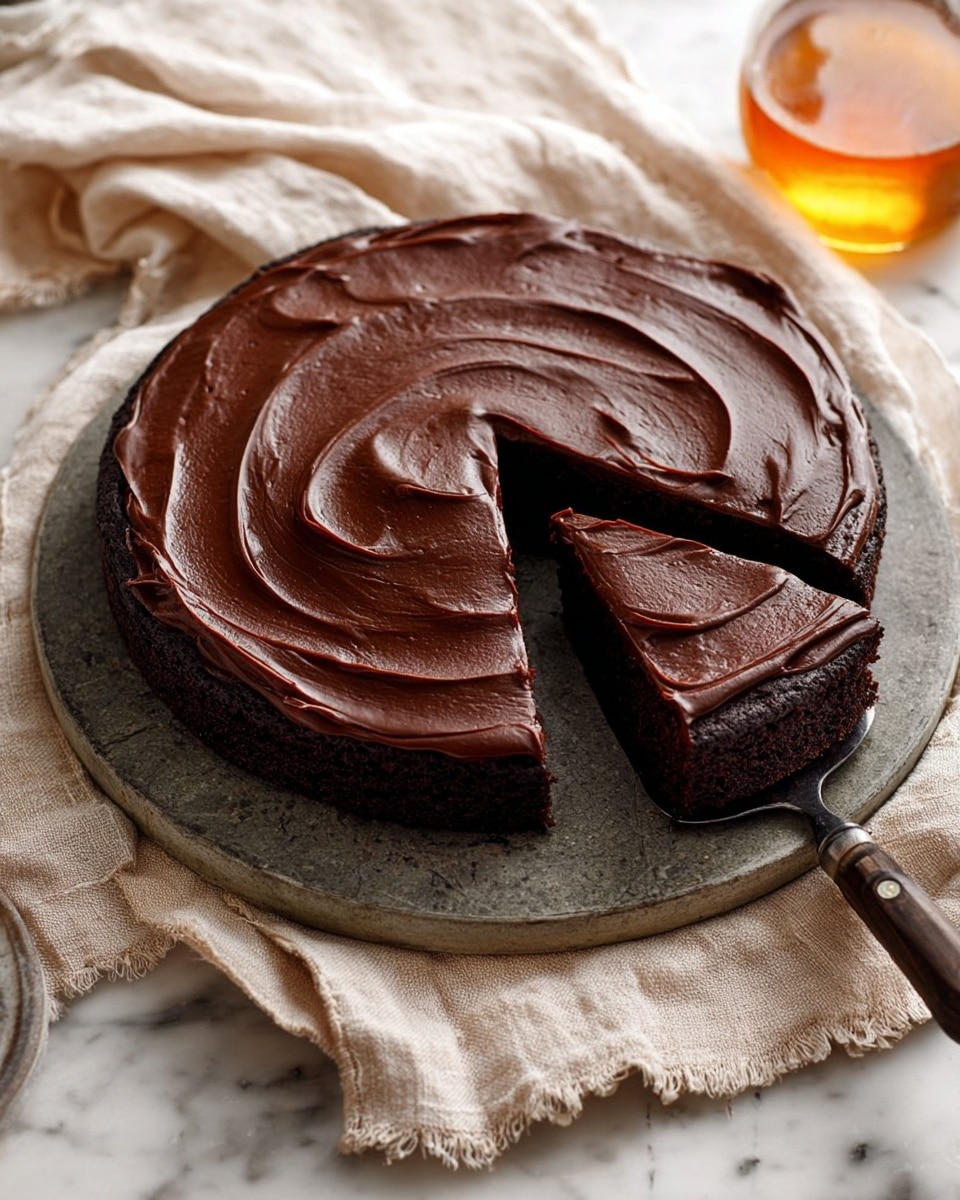 A dark chocolate cake with one smooth layer of thick, glossy chocolate frosting spread evenly on top, showing visible swirls and waves. The cake is round and sits on a round stone gray plate placed on a cream-colored cloth with a rustic texture. One triangular slice of the cake is lifted out with a kitchen spatula that has a dark wooden handle, revealing the dense, moist texture of the cake's inside, which matches the dark chocolate color of the frosting. In the background, there is a glass jug with a golden liquid on the cloth. The scene is set on a white marbled surface. photo taken with an iphone --ar 4:5 --v 7