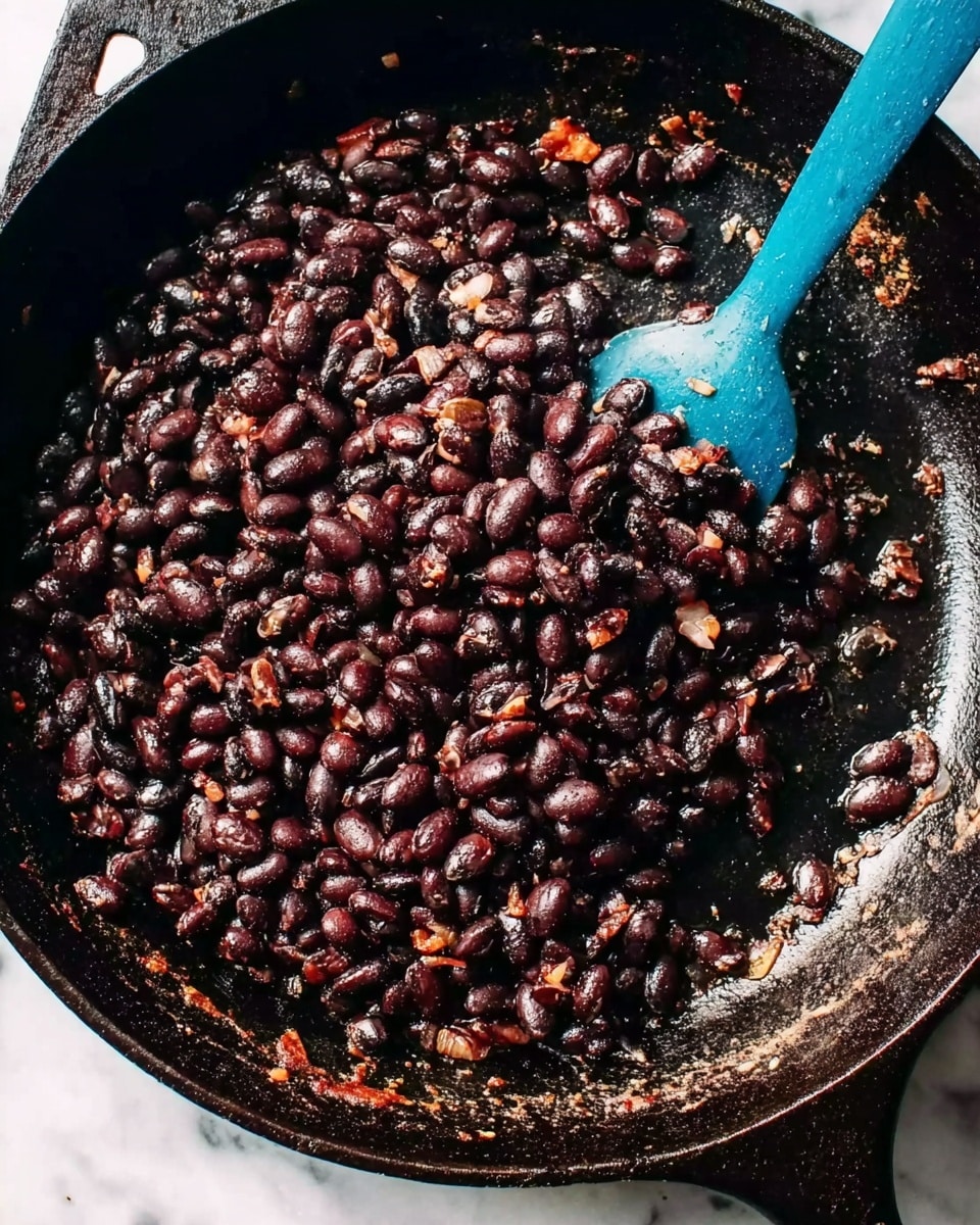 The image shows a close-up of a black cast iron pan filled with dark cooked black beans. The beans look soft and slightly shiny with bits of small, lighter brown pieces mixed in. A blue spatula is stirring the beans on the right side of the pan. The pan's surface shows some areas with a rusty or worn look, and the pan rests on a white marbled surface. The overall colors are deep brown and black with the bright blue spatula standing out. photo taken with an iphone --ar 4:5 --v 7