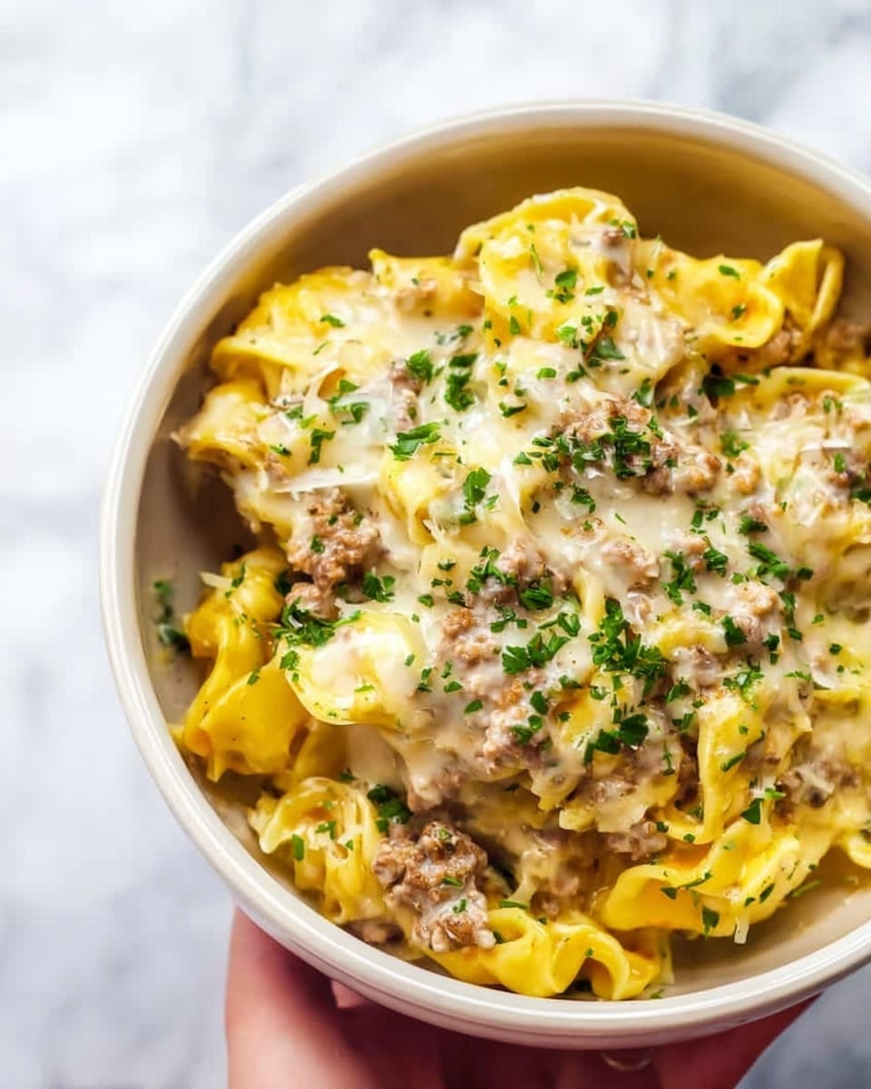 The dish is presented in a white bowl filled with a creamy mix of pasta and sauce. It has multiple layers, starting with small, yellow pasta pieces that look soft and slightly chewy. On top of the pasta, there is a creamy white cheese sauce spread evenly, melting around the pasta. Small bits of browned meat or ground meat are mixed throughout, adding a textured look. The dish is garnished with finely chopped green herbs scattered over the top, adding a fresh contrast to the cream and yellow colors. The bowl sits on a white marbled surface, and a woman's hand is holding it from the side. Photo taken with an iphone --ar 4:5 --v 7