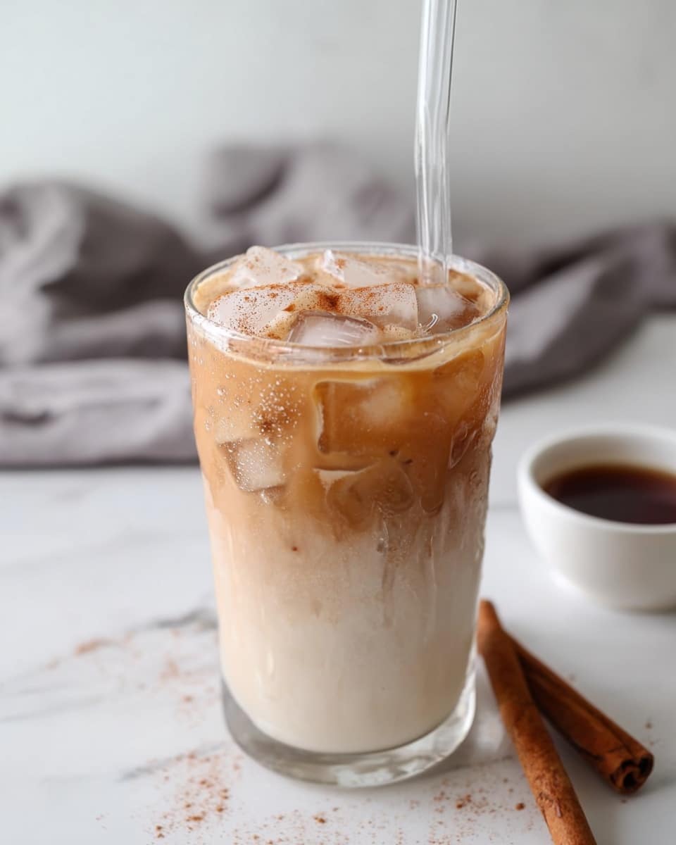 A clear glass filled with iced coffee shows three visible layers: at the bottom a creamy white layer, in the middle a light tan layer blending softly into the white, and a top layer of slightly darker brown coffee with ice cubes floating, some showing a frothy surface sprinkled lightly with cinnamon powder. A clear straw is placed in the glass. The glass sits on a white marbled surface with a cinnamon stick to the right and a small white bowl with dark syrup in the blurred background, along with a soft gray cloth. photo taken with an iphone --ar 4:5 --v 7