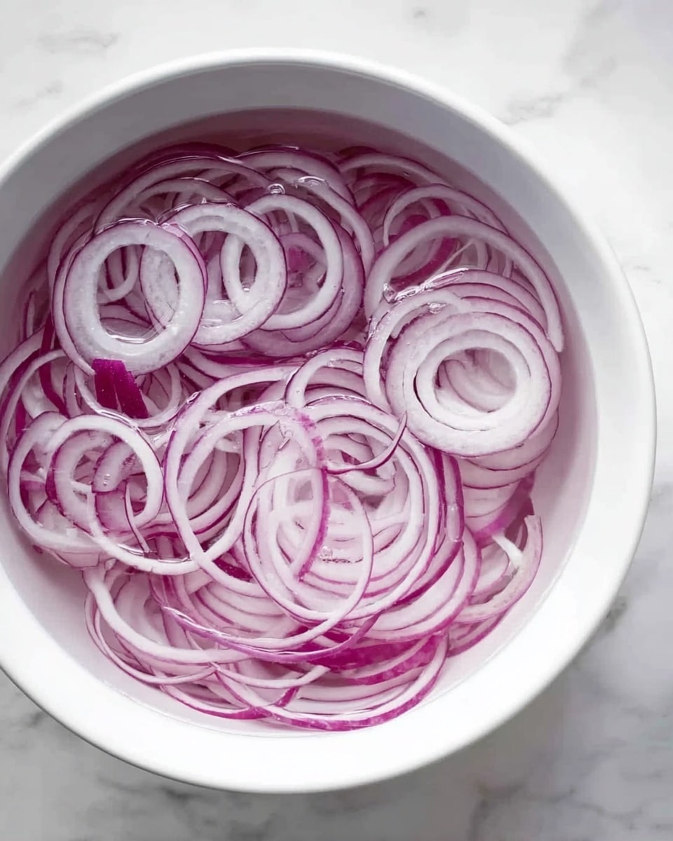 A white bowl filled with many thin rings of purple onion, soaking in clear water. The onion rings show layers with different shades of purple and white, some are curled and overlapping inside the bowl. The bowl is placed on a white marbled surface. photo taken with an iphone --ar 4:5 --v 7