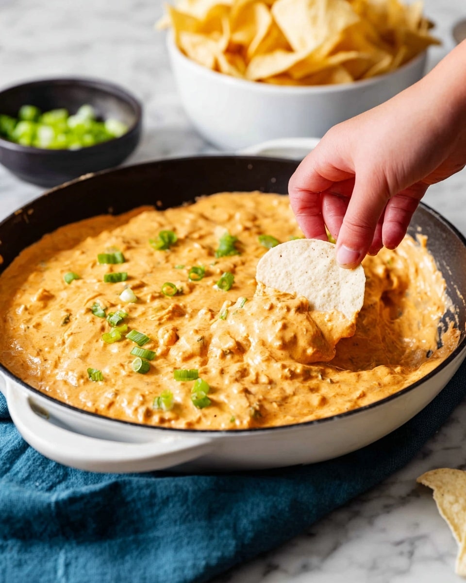 A round white skillet filled with a thick, creamy orange cheese dip that has a slightly chunky texture. The dip is sprinkled with small pieces of fresh green onions scattered on top, adding a touch of bright green color. A woman's hand is dipping a large, light beige tortilla chip partly coated with the cheese dip into the skillet from the right side of the image. In the background, a white bowl filled with golden tortilla chips is slightly out of focus, next to a small black bowl holding chopped green onions. The skillet rests on a folded dark blue cloth on a white marbled surface photo taken with an iphone --ar 4:5 --v 7