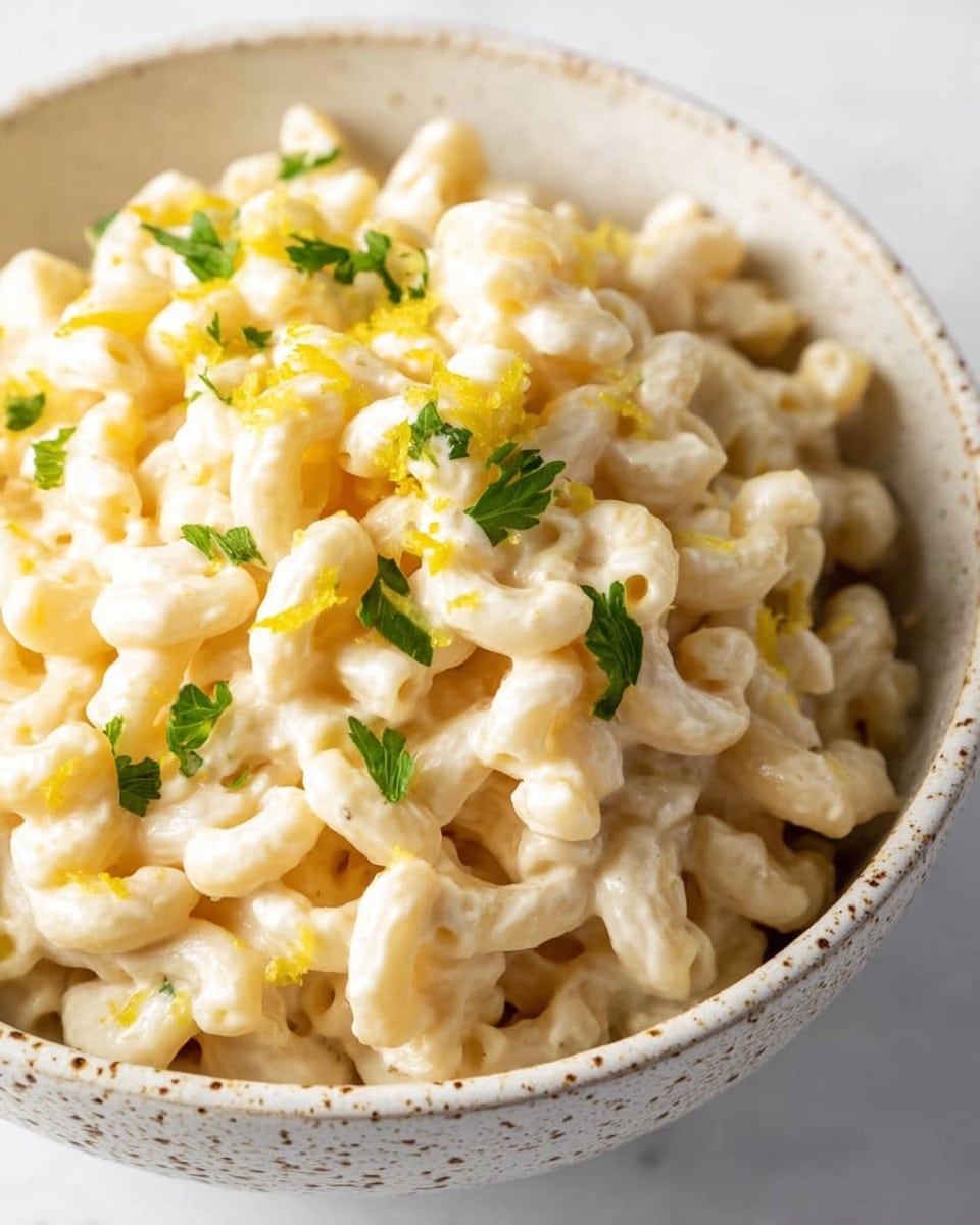 A close-up of a bowl filled with creamy macaroni. The macaroni is coated in a pale, smooth sauce with small herbs mixed in. On top, there are small green parsley leaves and light yellow lemon zest sprinkled to add color. The bowl is white with small brown speckles and is placed on a white marbled surface. Photo taken with an iphone --ar 4:5 --v 7