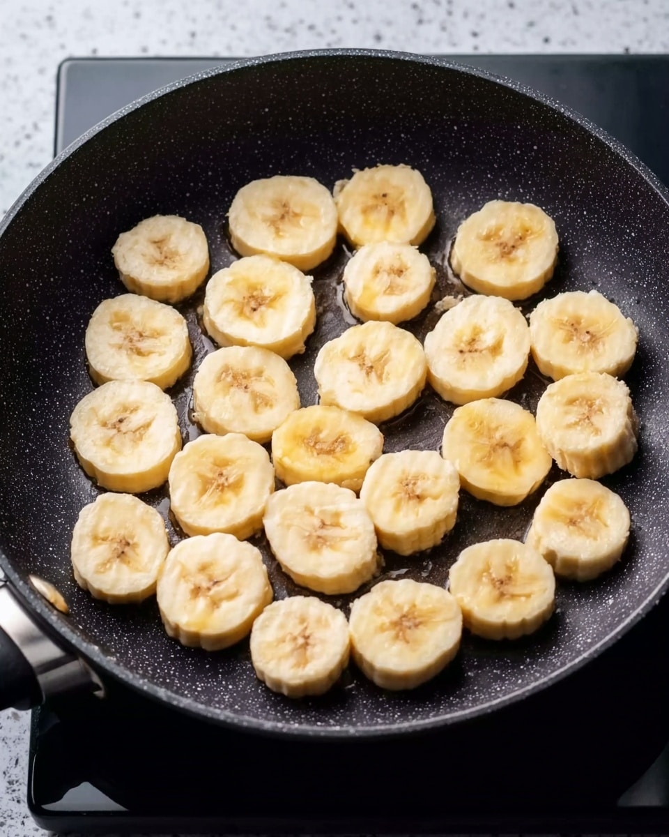 A black frying pan filled with evenly sliced banana pieces arranged in a single layer, each slice showing a soft yellow color with a pale center and slightly rounded edges. The pan sits on a stove with a faintly visible burner and a dark speckled surface replaced by a white marbled texture. The bananas have a smooth and slightly moist texture, beginning to brown gently on their flat surfaces as they cook. Photo taken with an iphone --ar 4:5 --v 7