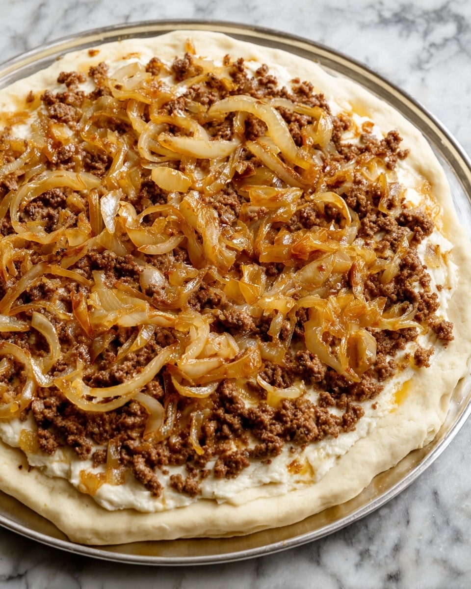 The image shows an uncooked pizza on a shiny silver tray placed on a white marbled surface. It has three main layers: the base layer is pale beige dough with a soft, slightly puffy edge; the middle layer is a spread of finely crumbled brown cooked meat covering the inner part of the dough; and the top layer consists of glossy, golden-brown caramelized onion slices scattered unevenly over the meat. The textures of the onions are smooth and slightly translucent, contrasting with the rough, crumbly meat beneath. Photo taken with an iphone --ar 4:5 --v 7