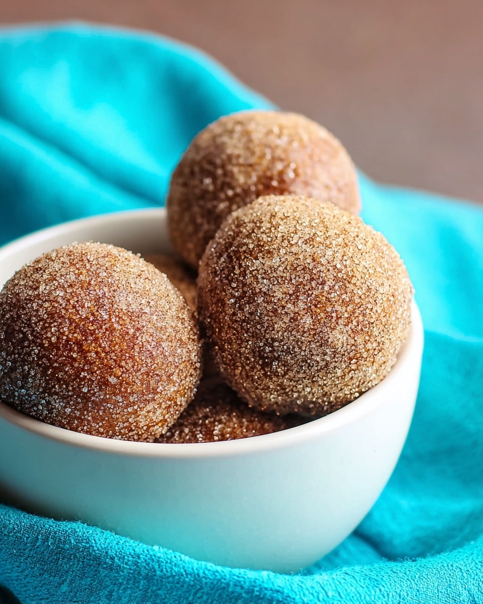 The image shows four round doughnut holes with a coarse sugar coating, giving a textured look with a light brown color. They are placed inside a white bowl, with one doughnut hole prominently in front and the others partially visible behind it. The bowl sits on a bright aqua cloth that is softly folded around it, with a background that transitions into a soft brown blur. The lighting highlights the sparkling sugar crystals on the doughnut holes, making them look fresh and tasty. photo taken with an iphone --ar 4:5 --v 7
