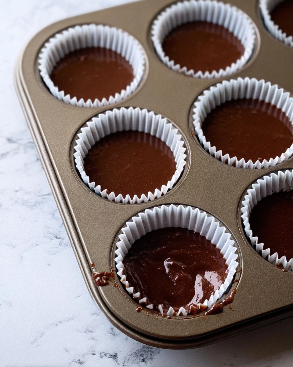 The image shows a metal muffin tray with six cups lined with white paper baking liners. Each liner is filled with smooth, dark brown chocolate batter spread evenly inside. One cup has some batter spilled on the edge and outside the liner. The muffin tray is placed on a table with a clean white marbled surface. The focus is on the texture of the chocolate batter and the neat paper liners. photo taken with an iphone --ar 4:5 --v 7