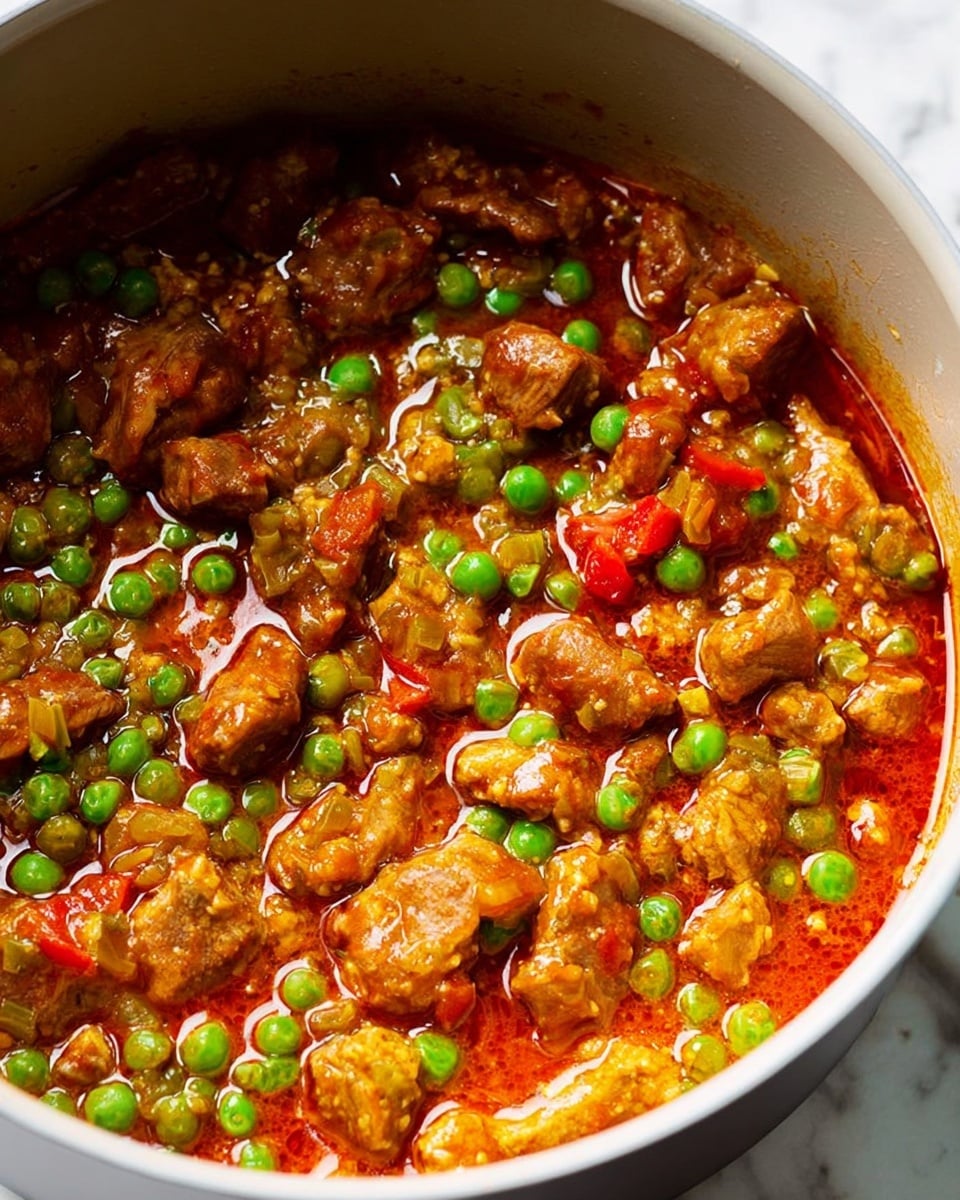 A close-up view of a saucepan filled with a stew made of small, brown, cooked meat pieces mixed with bright green peas and small red bell pepper chunks. The stew has a shiny, oily orange-red sauce with a slightly thick texture, covering the meat and vegetables. The saucepan is white on the inside, sitting on a white marbled textured surface. The focus is on the rich sauce and tender meat pieces floating in it, with the green peas adding a pop of color throughout the dish. photo taken with an iphone --ar 4:5 --v 7