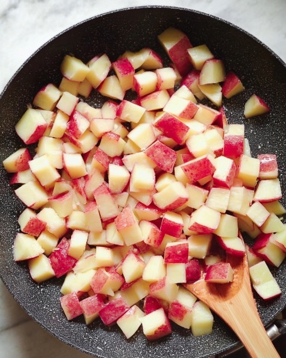 The image shows a black frying pan filled with many small cubes of potatoes with red skin. The potato cubes fill the pan evenly, showing a mix of creamy white inside and red skin edges. A wooden spoon is partly visible on the right side of the pan, stirring the potatoes. The pan sits on a white marbled surface. photo taken with an iphone --ar 4:5 --v 7