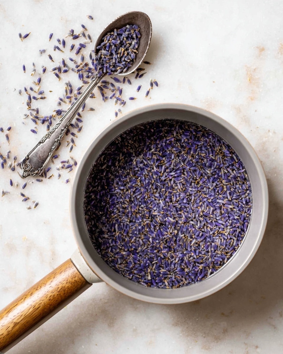 A light gray pot with a wooden handle is filled with water and a thick layer of small, dark purple dried lavender buds floating on the surface, creating a rich textured pattern that covers the entire top view of the water. Above the pot, an antique silver spoon lies on a white marbled surface, holding some scattered lavender buds. The whole scene is set against a soft white marbled background that highlights the deep purple hues and silver tones clearly. photo taken with an iphone --ar 4:5 --v 7