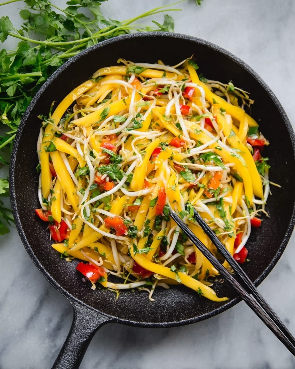 A black cast iron pan holds a colorful mango salad with three main layers: long, bright yellow mango strips on the bottom, topped with thin white bean sprouts, and sprinkled with small diced red bell peppers and chopped green herbs evenly mixed throughout. Two black chopsticks rest across the right side of the pan, slightly picking up some salad pieces. The scene is set on a white marbled surface with leafy green stems scattered to the left side. Photo taken with an iphone --ar 4:5 --v 7