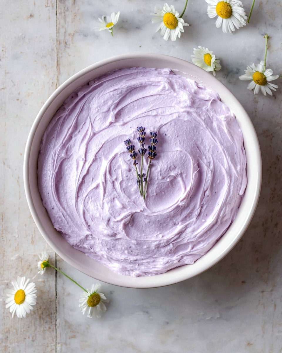 A single layer of light purple whipped cream with soft swirled textures fills a large white bowl, decorated simply with two small sprigs of lavender placed in the center. The bowl sits on a white marbled surface with a few small white and yellow daisy flowers casually spread around the edges. The overall look is fresh, smooth, and pastel-toned, with a natural, delicate decoration. photo taken with an iphone --ar 4:5 --v 7