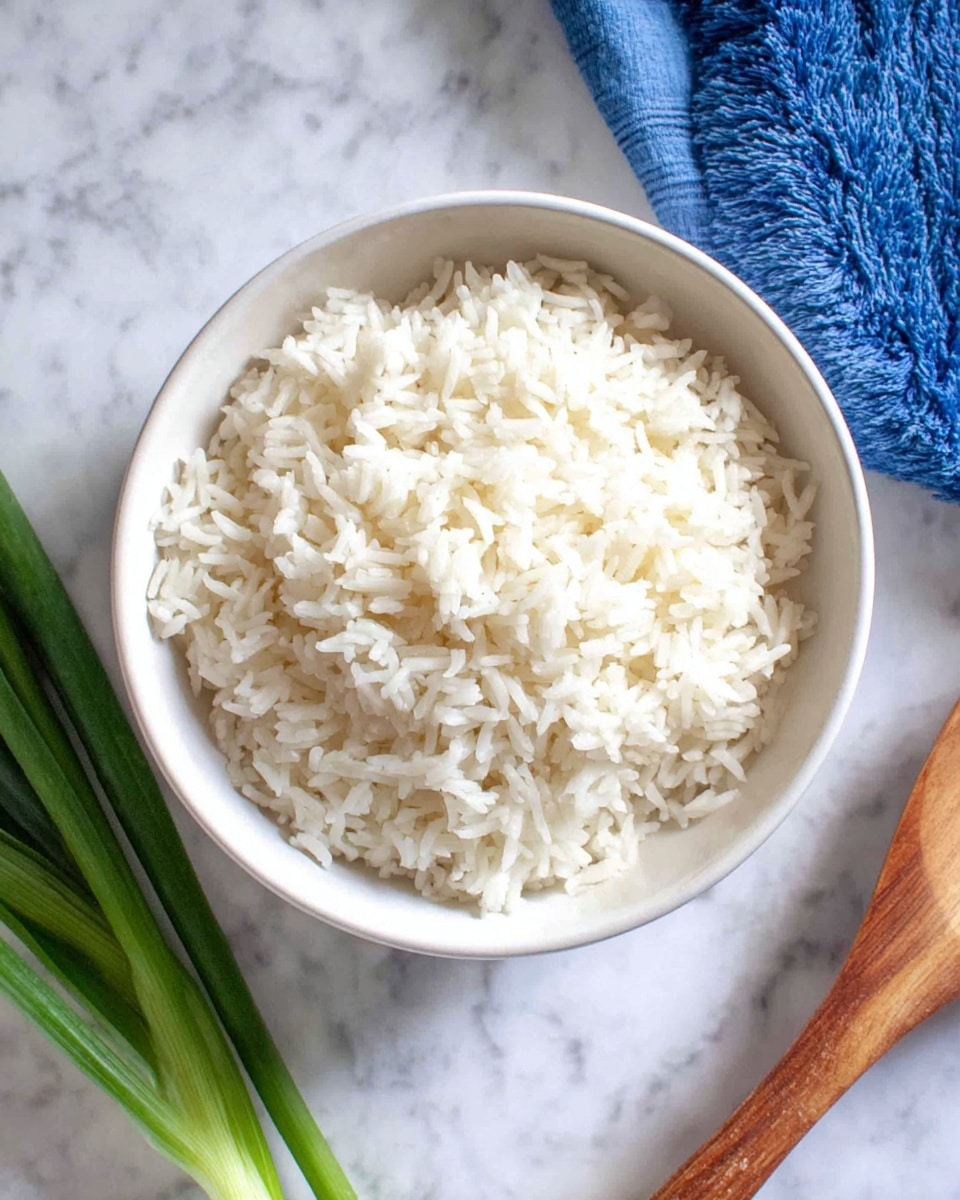A close-up top view of a white bowl filled with cooked white rice, showing soft, separate grains piled up evenly, placed on a white marbled surface. To the left, there are some green onions and a blue cloth partially visible, and on the right, part of a wooden spoon can be seen. The scene is bright and clean. photo taken with an iphone --ar 4:5 --v 7