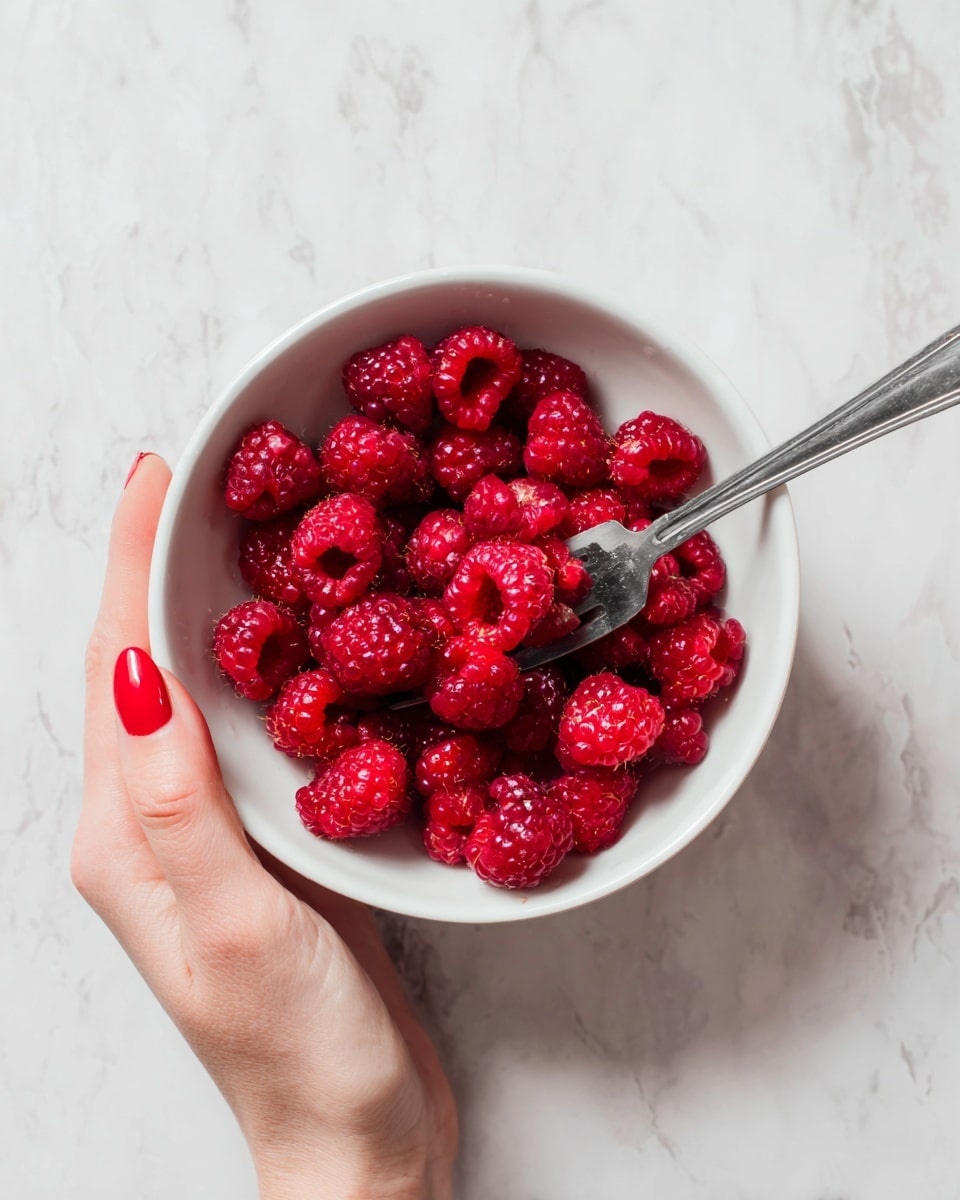 A white bowl filled with bright red raspberries, some of which look slightly softened and crushed. A woman's hand with red nail polish is holding a silver fork inside the bowl, pressing into the raspberries, while the other woman's hand is steadying the bowl from the side. The background and surface are a clean white marbled texture. photo taken with an iphone --ar 4:5 --v 7