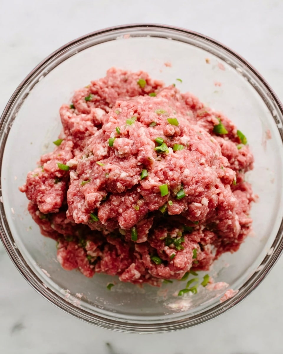 A clear glass bowl holds a mound of raw ground meat mixed with small green onion pieces scattered throughout. The meat is pinkish-red with a slightly coarse texture, showing the mix of ingredients within. The bowl is placed on a white marbled surface, and the image is taken from above, focusing on the contents inside the bowl. photo taken with an iphone --ar 4:5 --v 7