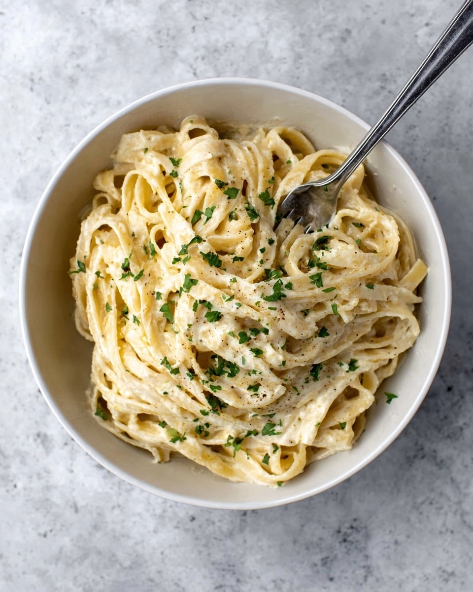 A white bowl filled with creamy fettuccine pasta showing about three thick layers of noodles coated in a smooth, pale yellow sauce. The pasta is sprinkled lightly with chopped green herbs, adding a touch of color on top. A silver fork rests inside the bowl, slightly tangled in the pasta. The bowl is placed on a white marbled surface. photo taken with an iphone --ar 4:5 --v 7