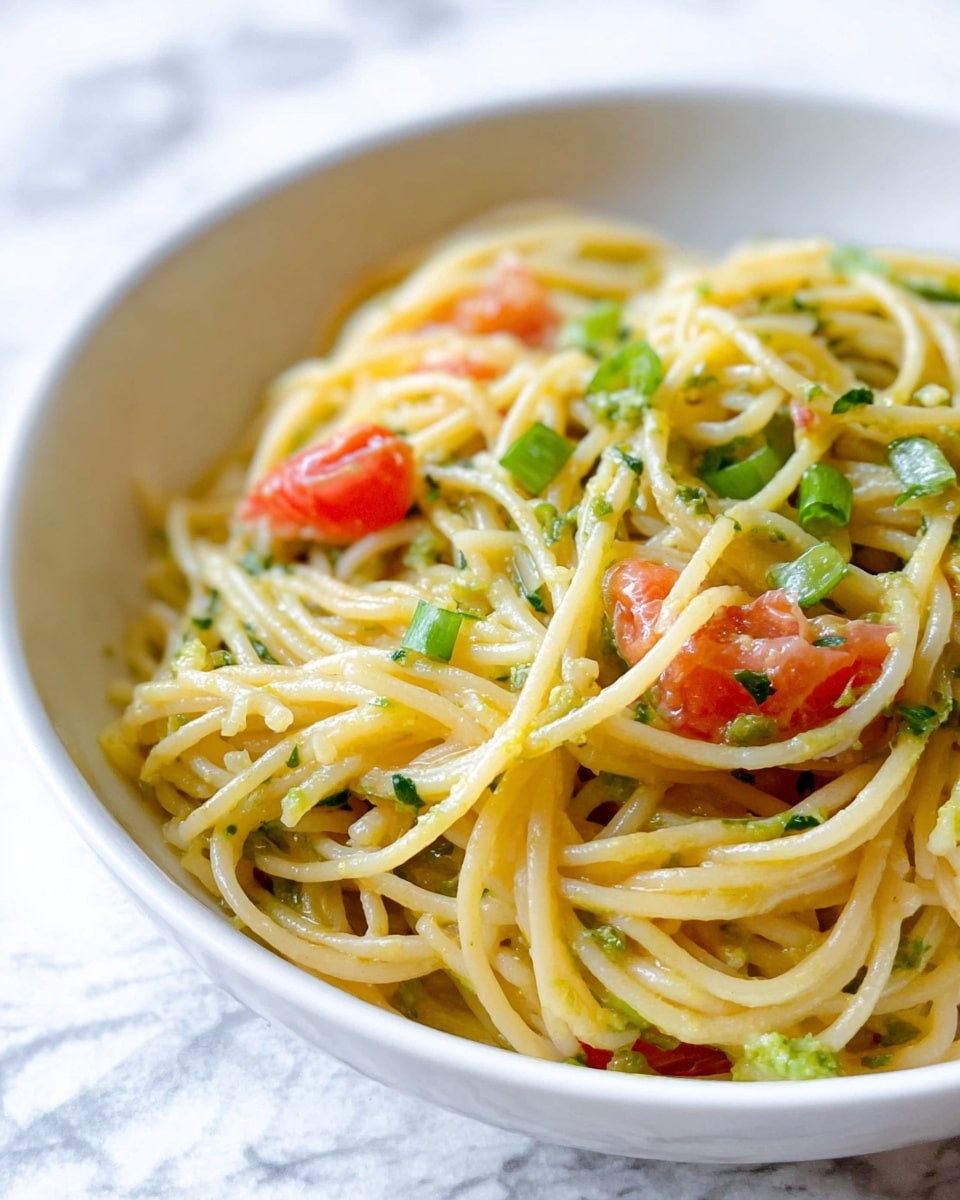 A white bowl holds a tangle of light yellow spaghetti noodles mixed with small pieces of green herbs and bright red tomato chunks. The noodles look soft and slightly shiny with a hint of sauce coating them. Scattered on top are small bits of fresh green onion adding texture and a pop of color. The bowl sits on a white marbled surface, creating a clean and fresh background for the dish. photo taken with an iphone --ar 4:5 --v 7