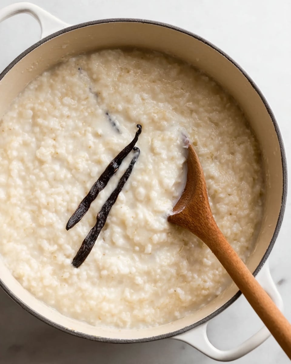 The image shows a close-up of a white pot filled with thick, creamy rice pudding. The pudding is light beige in color with a soft, slightly lumpy texture. Inside, there are two dark brown vanilla pods partially submerged near the center. A wooden spoon with a smooth surface is resting in the pudding on the right side of the pot. The pot is sitting on a white marbled surface. Photo taken with an iphone --ar 4:5 --v 7