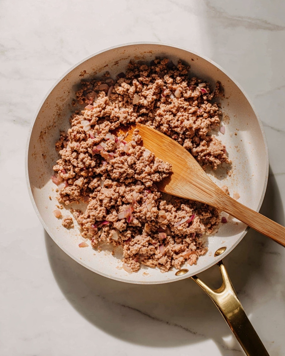 A white frying pan with a gold handle holds a mix of cooked ground meat and finely chopped onions. The meat is light brown with small pieces of reddish and translucent onion spread evenly throughout. A wooden spatula rests inside the pan, partly covered by the food, showing its smooth texture and natural wood color. The pan is placed on a white marbled surface with soft natural light creating gentle shadows around the edges. photo taken with an iphone --ar 4:5 --v 7