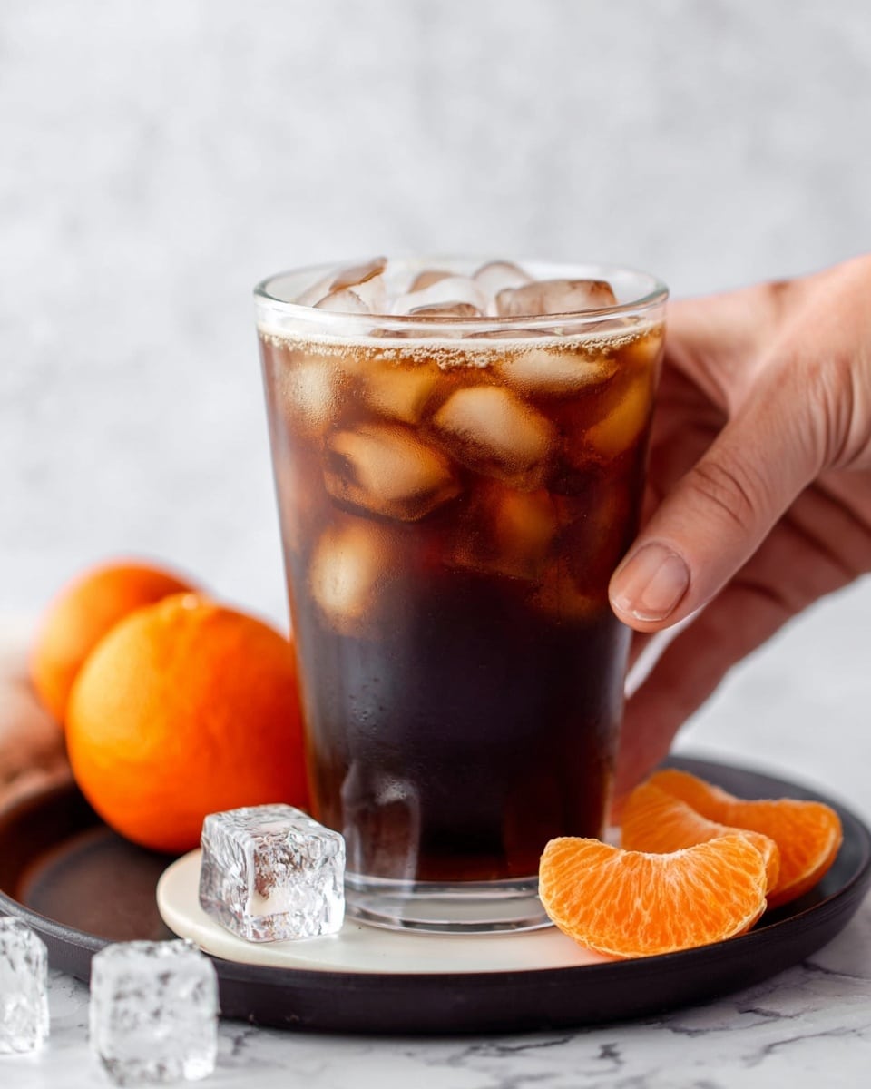 A clear glass filled with dark brown cold coffee, layered with many large, translucent ice cubes on top, creating a frosty texture near the rim. The glass is placed on a white round coaster over a black tray. In the foreground, three bright orange tangerine slices and two clear ice cubes rest on the tray. A woman's hand with visible skin texture is gently holding the glass from the right side. The surface beneath everything is a white marbled texture. photo taken with an iphone --ar 4:5 --v 7