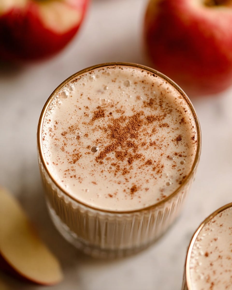 The image shows a close-up top view of a creamy drink in a clear glass with a gold rim, filled with a light beige liquid that has tiny specks and a dusting of brown powder on the surface. The glass is placed on a white marbled surface. Around the glass, there is a blurred red apple and another glass of the same drink mostly out of focus. The drink looks smooth and frothy with small bubbles on top. Photo taken with an iphone --ar 4:5 --v 7
