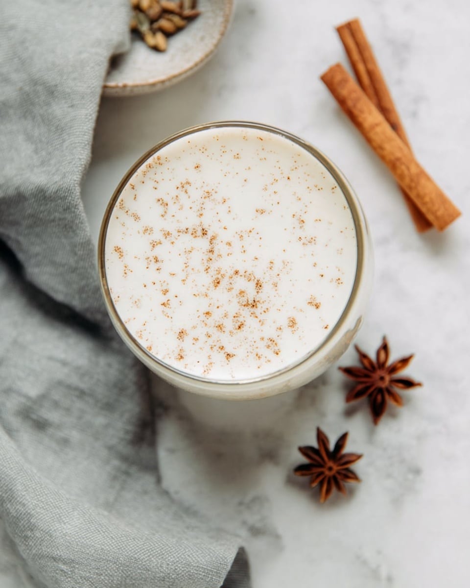 A glass cup filled with a creamy white drink topped with small brown spice specks, placed on a white marbled surface. Nearby, there is a whole cinnamon stick and two star anise pods adding a warm brown contrast. A soft gray cloth is gently folded to the side, complementing the calm tones in the scene. The top view shows the smooth texture of the drink with tiny speckles evenly spread across. photo taken with an iphone --ar 4:5 --v 7