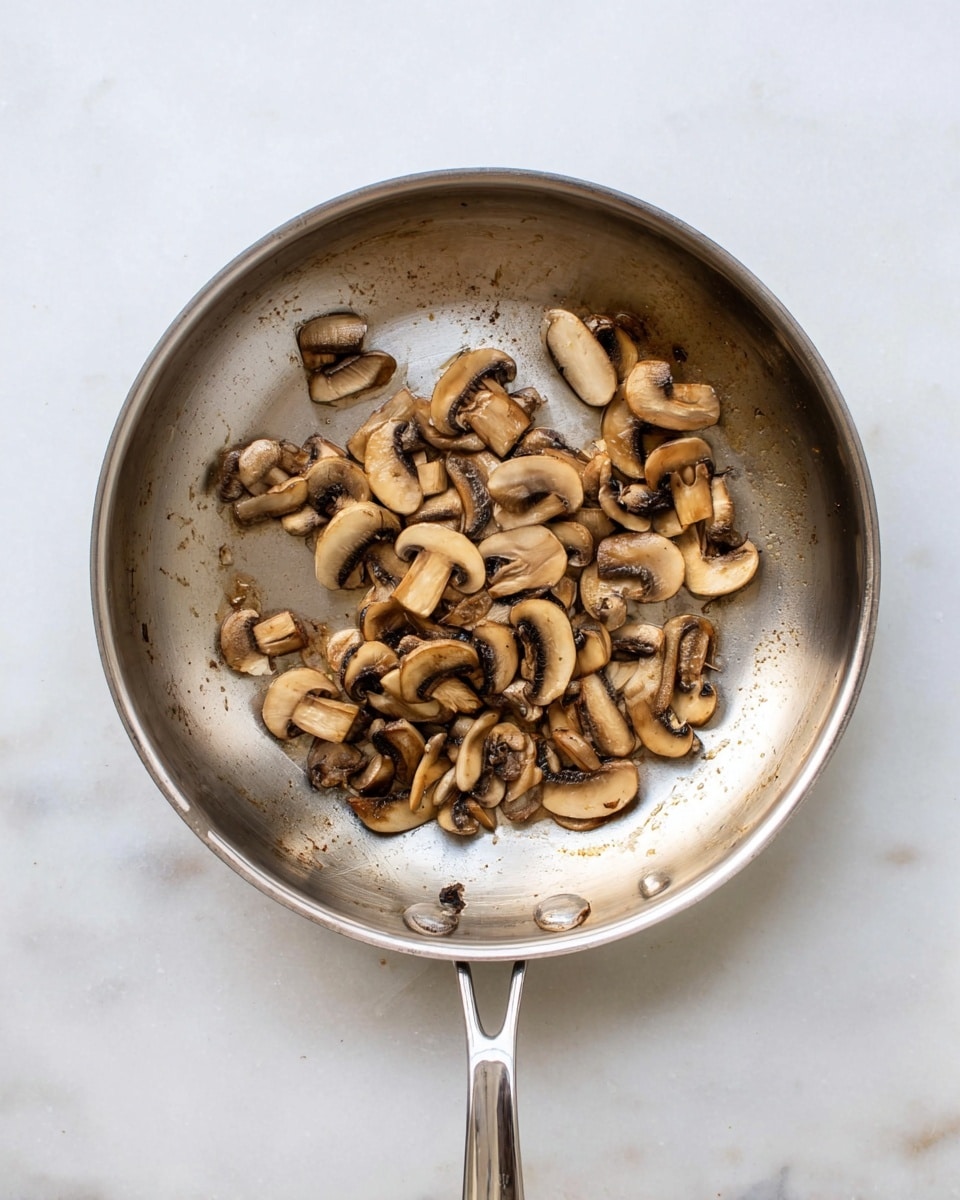 A round silver pan sits on a white marbled surface, filled with a single layer of sliced cooked mushrooms spread unevenly, mostly gathered on one side of the pan. The mushrooms are light to dark brown with a soft, moist texture and show some slight browning from cooking. The pan handle is visible at the top and bottom, and the pan interior shows slight signs of use with oil spots. photo taken with an iphone --ar 4:5 --v 7
