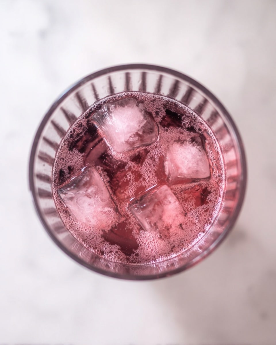 Top view of a tall clear glass filled with a pink bubbly liquid and several large ice cubes floating inside. The glass has vertical ridges on the sides, and the drink shows tiny bubbles on the surface. The glass is placed on a white marbled surface. photo taken with an iphone --ar 4:5 --v 7