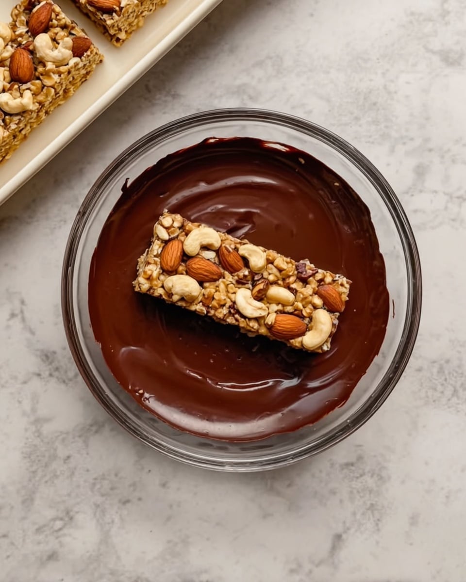 The image shows a clear glass bowl placed on a white marbled surface. Inside the bowl, there is a layer of smooth, melted dark chocolate covering the bottom, with a nut and seed bar resting on top in the center. The bar has one visible layer packed with mixed nuts like almonds, cashews, and peanuts, all light brown and beige in color with a crunchy texture. In the upper left corner, part of a white tray with more nut and seed bars is slightly visible. The setting looks like a kitchen scene focused on dipping or coating the bar with chocolate. photo taken with an iphone --ar 4:5 --v 7