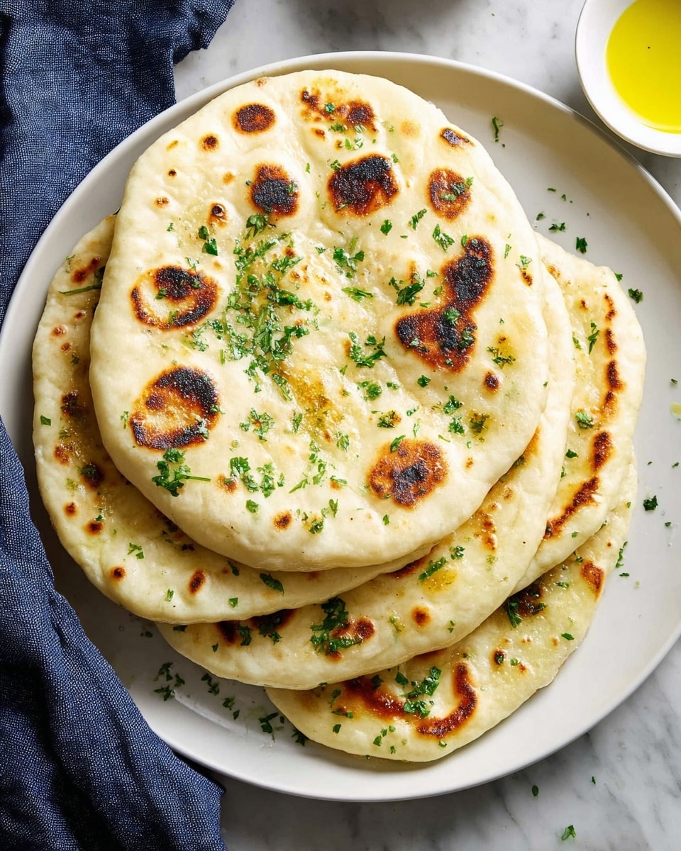 A white plate holds a stack of five flatbreads. Each flatbread is round and light golden with brown charred spots on top. Bright green chopped herbs are sprinkled over the flatbreads and scattered around the plate. The flatbreads have a soft, slightly puffy texture with uneven edges. The plate sits on a white marbled surface with a dark blue cloth visible on the left side. Part of a white bowl with yellow liquid is in the top right corner. photo taken with an iphone --ar 4:5 --v 7