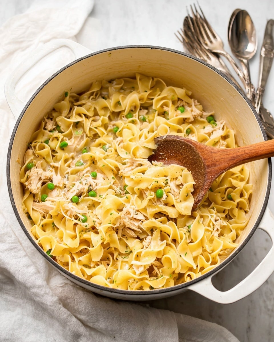 In a large white pot, there is one main layer of yellow, curly egg noodles mixed with small bits of light brown shredded chicken and scattered green peas. A wooden spoon is partly inside the pot, lifting some noodles, showing their soft and slightly shiny texture. The pot sits on a white marbled surface near silver forks and a white cloth napkin in a casual setting. The colors are warm and natural, with a cozy, home-cooked feel. photo taken with an iphone --ar 4:5 --v 7