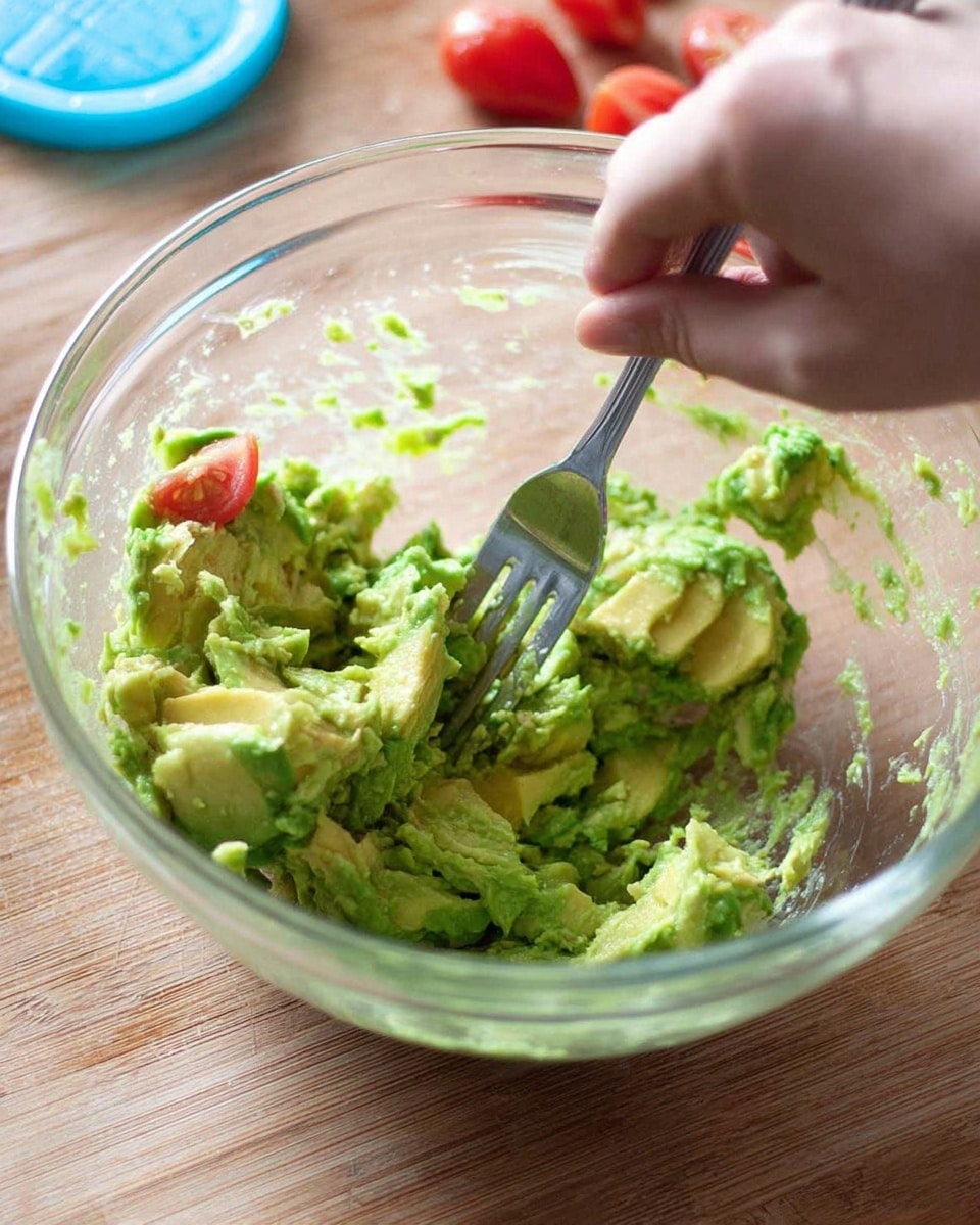 A woman's hand is using a silver fork to mash bright green avocado pieces inside a clear glass bowl. The avocado is unevenly smashed, showing chunks and smoother mashed parts. In the background on a wooden surface, there are small pieces of red tomato near a blue plastic lid. The texture of the avocado is creamy with some visible rough and soft spots. Photo taken with an iphone --ar 4:5 --v 7