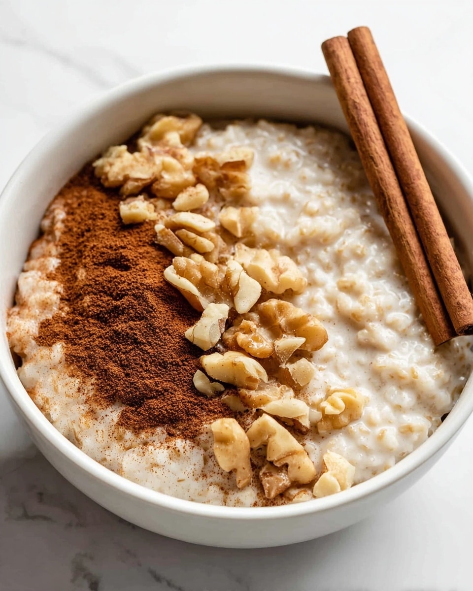 A white bowl filled with creamy, textured rice pudding forms the base layer covering most of the bowl's right side. On the left side, a layer of reddish-brown ground cinnamon powder spreads in a neat half-moon shape. Overlapping partially in the middle, golden-brown chopped walnut pieces are scattered, creating a rough and crunchy texture. Two whole cinnamon sticks lie diagonally on top of the cinnamon powder and walnuts, adding a smooth, woody look with warm brown tones. The bowl sits on a white marbled surface. Photo taken with an iphone --ar 4:5 --v 7