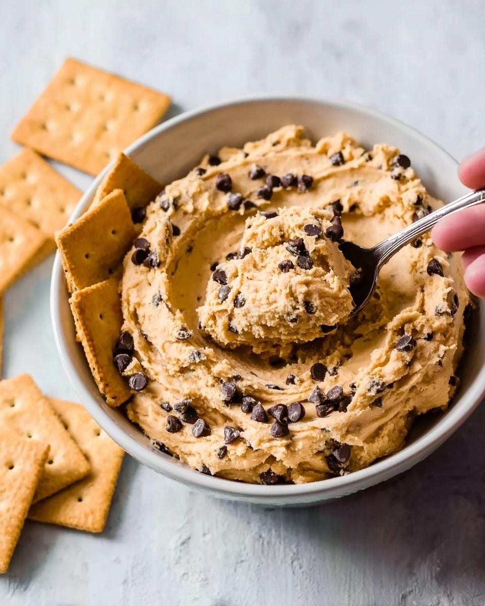 The image shows a white bowl filled with a light brown, creamy cookie dough with dark chocolate chips mixed throughout. Several square crackers are placed around the bowl's edge, partially inside the bowl. A woman's hand is holding a spoon scooping some of the cookie dough out. The bowl sits on a white marbled surface with a subtle texture, giving a clean and bright look. photo taken with an iphone --ar 4:5 --v 7