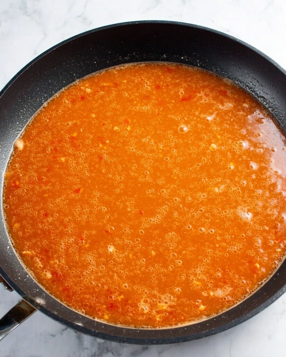 A black frying pan filled with a bubbling orange sauce with small pieces of ingredients visible throughout, sitting on a white marbled surface. The sauce has a smooth texture with some bubbles and slight patches of oil on the top, covering the entire pan evenly. Photo taken with an iphone --ar 4:5 --v 7