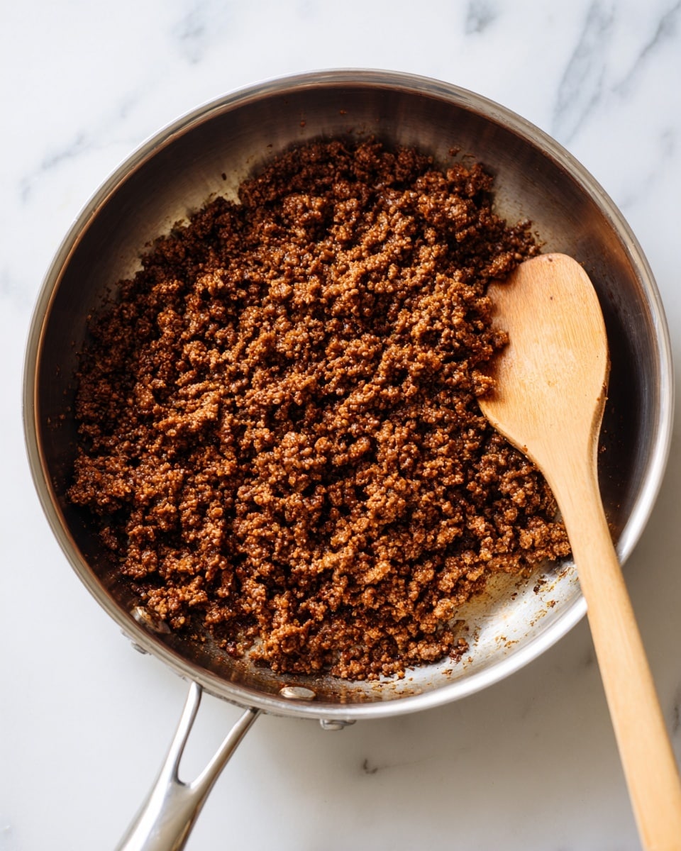 A close-up view of a large silver metal frying pan filled with one layer of small, crumbly brown cooked ground meat covering most of the pan's surface. On the right side of the pan, there is a wooden spoon with a smooth texture resting on top of the meat. The pan is placed on a white marbled surface, visible around its edges. Photo taken with an iphone --ar 4:5 --v 7