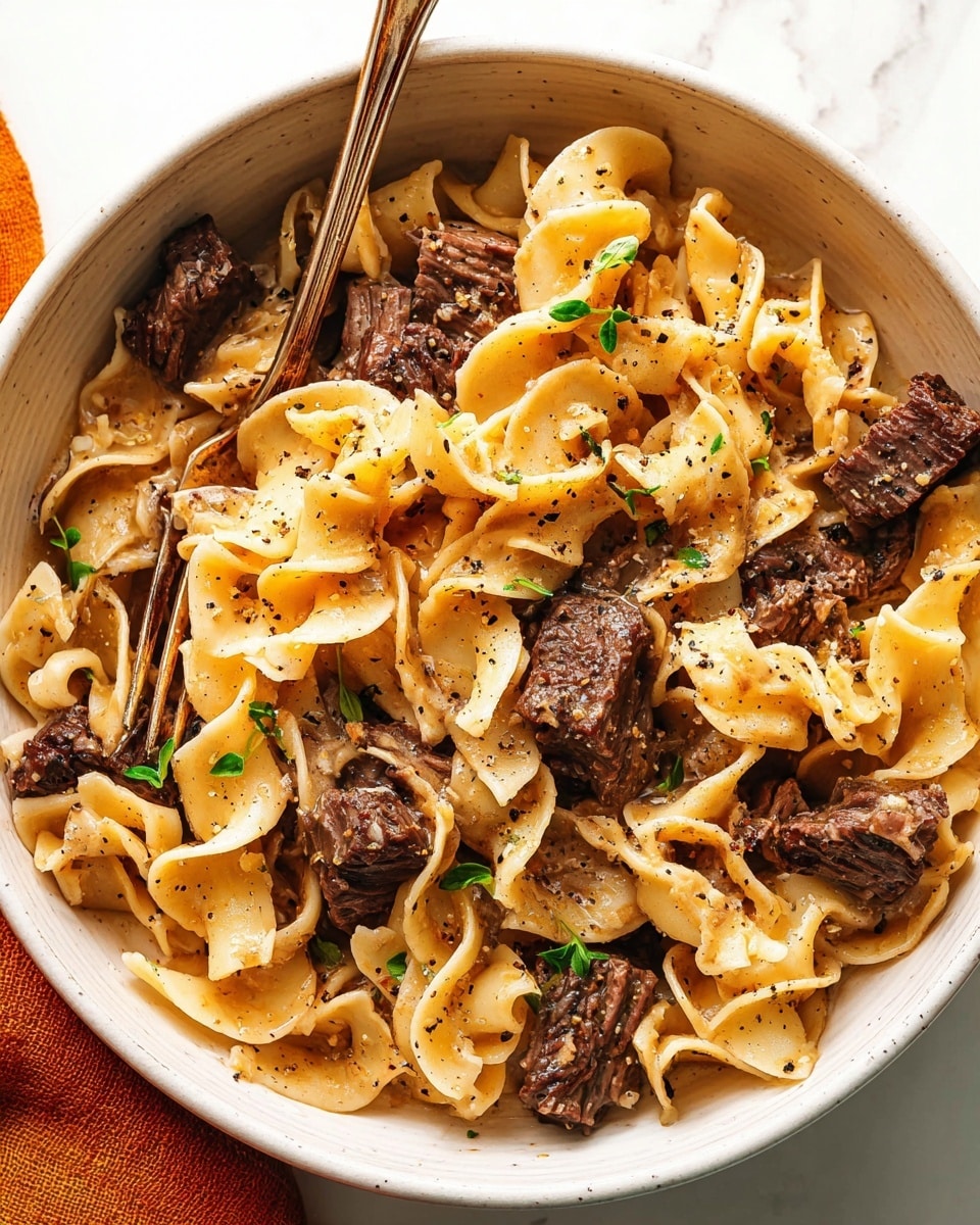 A close-up view of a white bowl filled with wide, curly egg noodles layered with chunks of dark brown, tender beef scattered evenly throughout. The noodles are coated in a light brown sauce and garnished with small green herb leaves. Freshly cracked black pepper is sprinkled on top, adding texture and contrast. A metal fork is partially placed inside the bowl on the left side, resting among the noodles and meat. The bowl sits on a white marbled surface with a hint of an orange cloth at the bottom left corner. Photo taken with an iphone --ar 4:5 --v 7