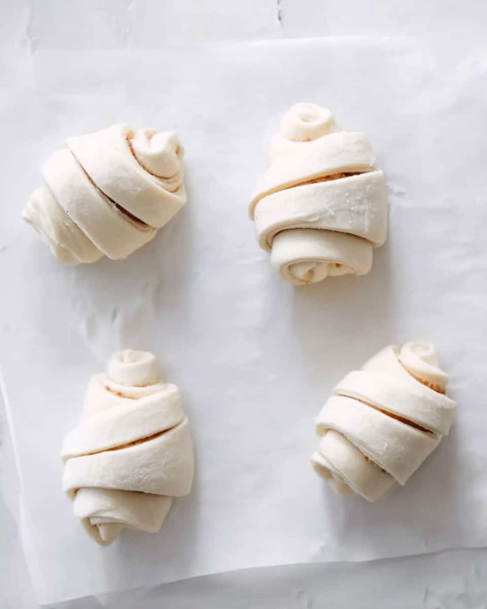 The image shows four small rolled dough pieces placed on white parchment paper over a white marbled surface. Each dough roll is made with many thin, smooth layers spiraled tightly to form a small cylinder shape, with light cream color dough and darker thin lines peeking through the edges, suggesting an inner filling. The dough pieces are evenly spaced in a loose grid, with soft shadows under each roll. The overall look is clean and simple. Photo taken with an iphone --ar 4:5 --v 7