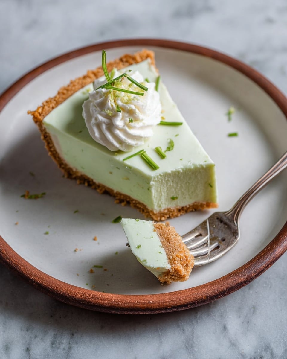 A slice of light green pie with two visible layers sits on a white plate with a brown rim; the bottom layer is a crumbly golden crust, and the top layer is smooth and pale green. On top of the pie slice is a small dollop of white whipped cream with thin, green herb pieces scattered around it and on the pie. A silver fork holds a small square piece of the pie, showing the two layers clearly. The plate is set on a white marbled surface. photo taken with an iphone --ar 4:5 --v 7